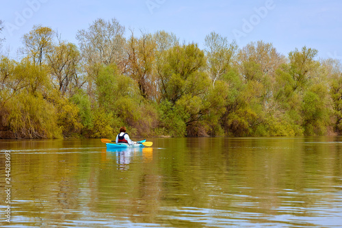 Rear view of woman paddle blue kayak on Danube river against the background of light green spring tree. Spring kayaking. Concept for adventure, travel, action, lifestyle.