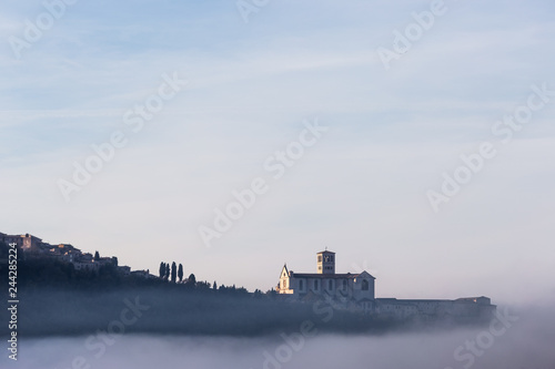 A view of St.Francis church in Assisi in the middle of mist beneath a blue sky with clouds © Massimo