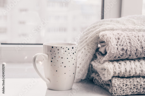Stack of warm cozy knitwears and a cup of coffee on white marble windowsill against white window background. Copy space.