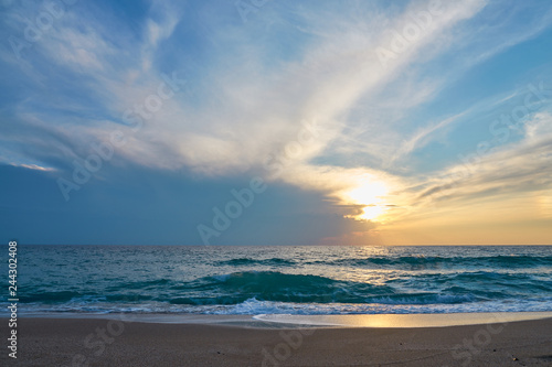 Sunset at the tropical beach, sun behind clouds reflects on water and waves with foam hitting sand.