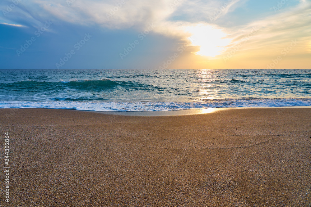 Obraz premium Sunset at the tropical beach, sun behind clouds reflects on water and waves with foam hitting sand.