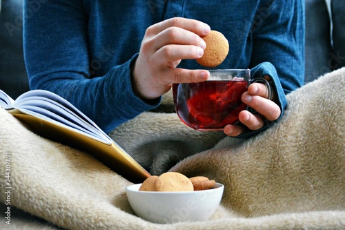 A young man is sitting on a sofa and reading a book while holding a fruit tea in a glass cup in hand and biscuits stand beside him - closeup on the book and glass cup full of tea - concept to warm up 