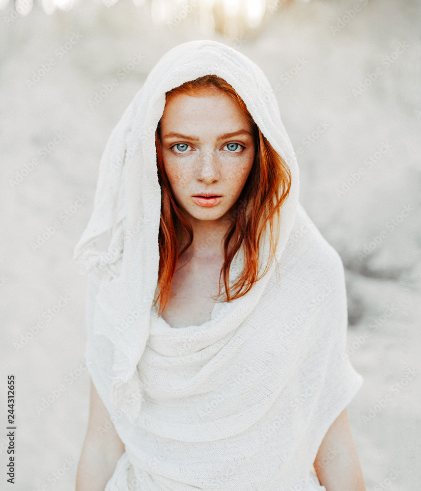 A beautiful young girl with red hair and freckles looks intently at the camera. Woman in the hood and clothes for the desert. Concept - obrazy, fototapety, plakaty 