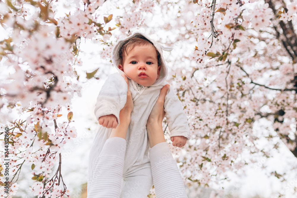 Portrait of happy joyful child in white clothes over tree flowers blossom background. Family playing together outside. Mom cheerfully hold little daughter Portrait of happy joyful child in white clothes over tree flowers blossom background. Family playing together outside. Mom cheerfully hold little daughter