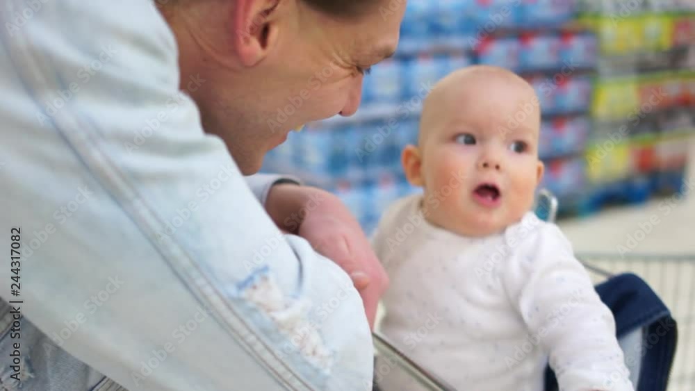Baby rides in a trolley in the supermarket with his father. The child carefully looks around Baby rides in a trolley in the supermarket with his father. The child carefully looks around