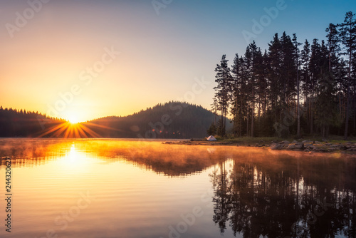 Sunrise on the lake Beautiful sunrise view with sun rays over a mountain lake surrounded by conifers. Shiroka Polyana dam in Rhodopi Mountains, Bulgaria.