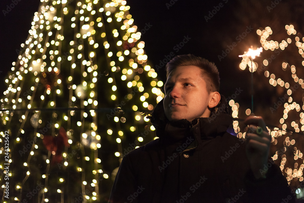 Dreamy handsome man standing on the street with sparkler in hand. Bright bokeh lights of Christmas tree and garlands on background. Winter holidays mood. Dreamy handsome man standing on the street with sparkler in hand. Bright bokeh lights of Christmas tree and garlands on background. Winter holidays mood.