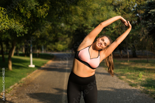 Pretty smiling plus size girl in pink sporty top and leggings happily looking in camera while stretching in cozy city park