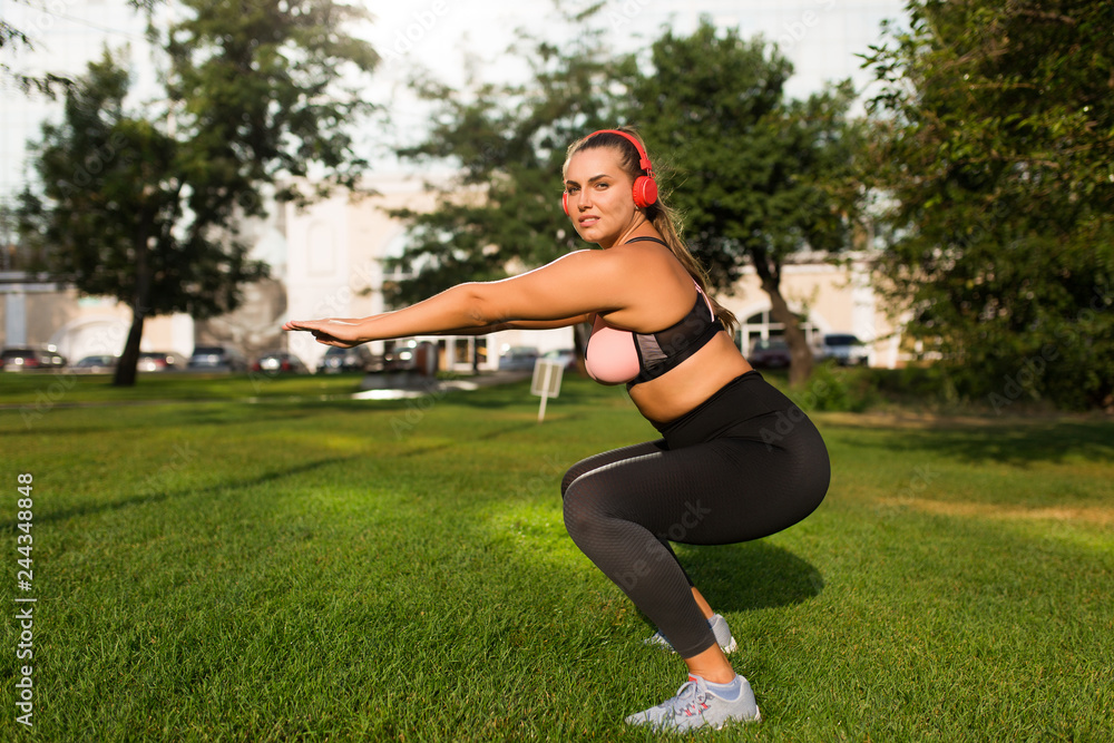 Young beautiful plus size woman in sporty top and leggings with red headphones dreamily looking in camera while doing sport exercises on grass in city park