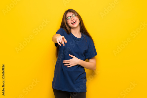 Young woman with glasses over yellow wall pointing with finger at someone and laughing a lot © luismolinero