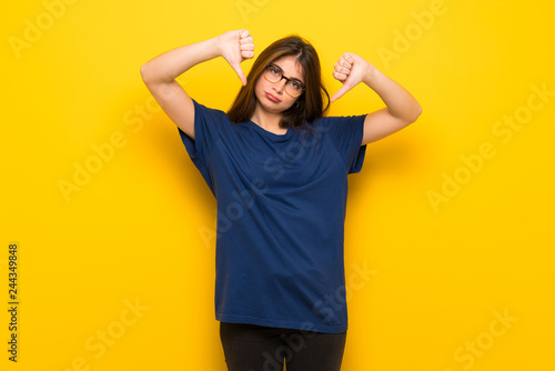 Young woman with glasses over yellow wall showing thumb down with both hands © luismolinero