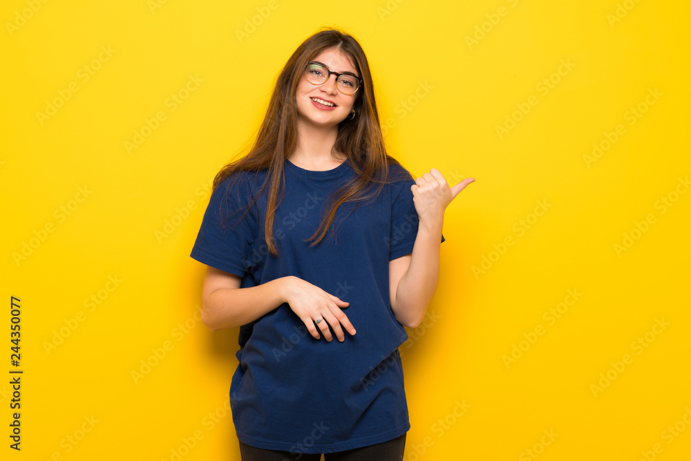 Young woman with glasses over yellow wall pointing to the side to present a product Young woman with glasses over yellow wall pointing to the side to present a product