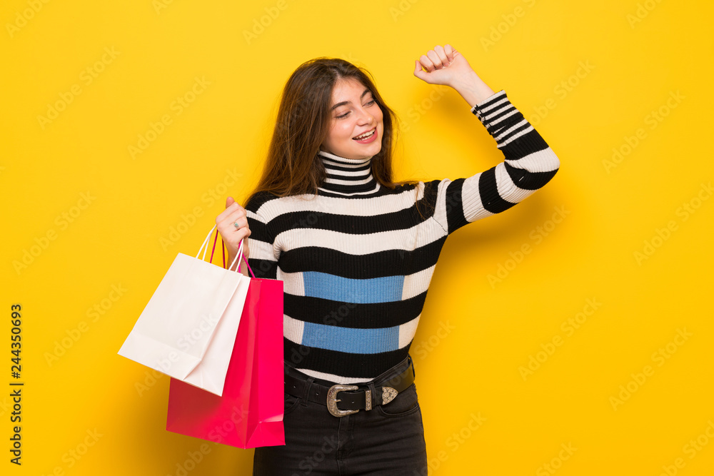 Young woman over yellow wall holding a lot of shopping bags in victory position Young woman over yellow wall holding a lot of shopping bags in victory position