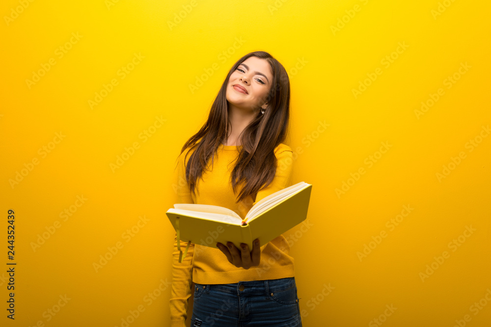 Teenager girl on vibrant yellow background holding a book and giving it to someone Teenager girl on vibrant yellow background holding a book and giving it to someone