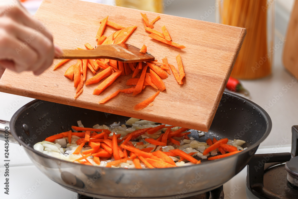 Home kitchen. A woman with a wooden cutting board adds carrots to a hot frying pan with vegetable oil and onions. Close-up.