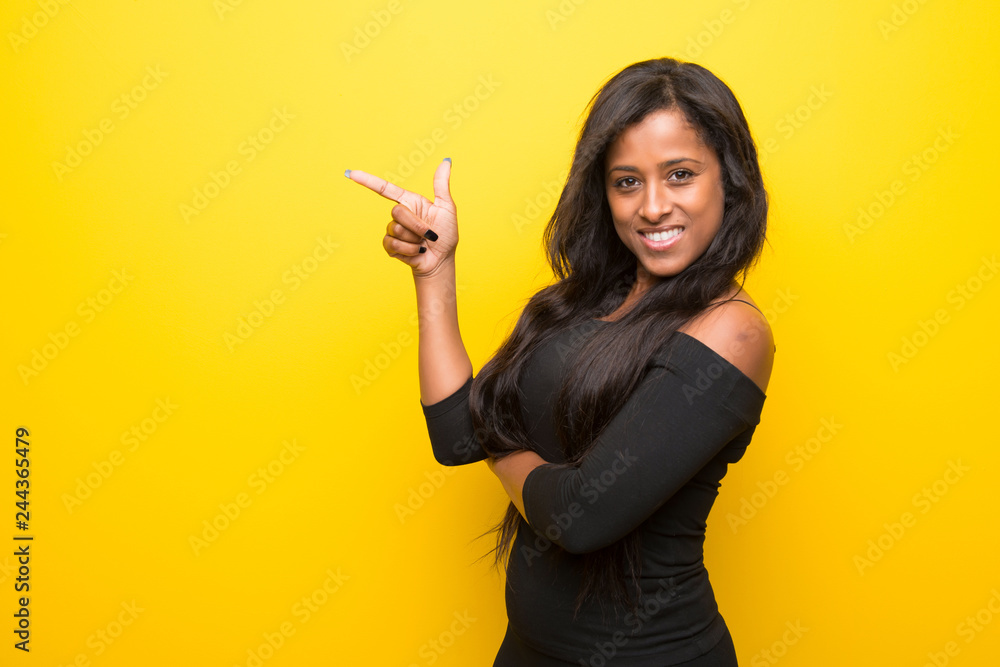 Young afro american woman on vibrant yellow background pointing finger to the side in lateral position Young afro american woman on vibrant yellow background pointing finger to the side in lateral position
