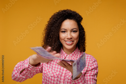 Portrait of an excited young african holding money banknotes and celebrating isolated over yellow background
