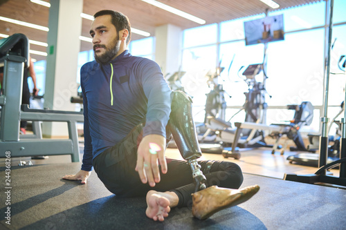 Bearded athlete with amputee leg sitting on the floor and resting in health club photo