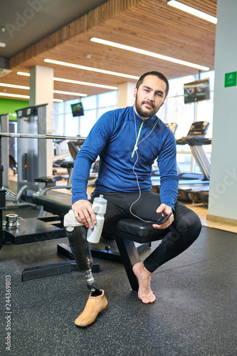 Portrait of bearded disabled athlete listening to music on smartphone and drinking water in fitness club