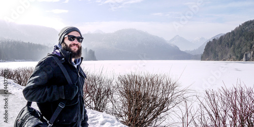 Man with beard in winter clothes looking out on a frozen lake and snowy distant mountains. Taken in Bavaria in winter.
