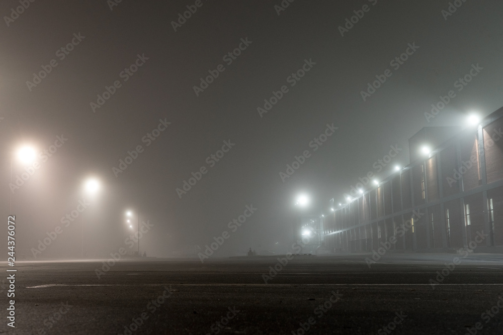 Fototapeta premium Empty urban car parking and streetlights at foggy night. Old Industrial brick building and lanterns on lonely street