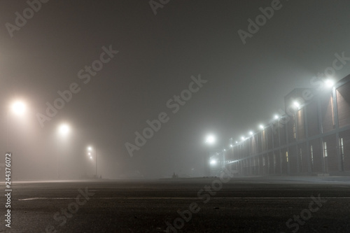 Empty urban car parking and streetlights at foggy night. Old Industrial brick building and lanterns on lonely street