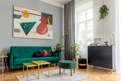 Luxury and modern home interior with design green sofa, navy commode, tables, pouf and accessroies. A lot of plants in the room. Abstract painting.  Stylish decor of living room with brown parquet.  photo