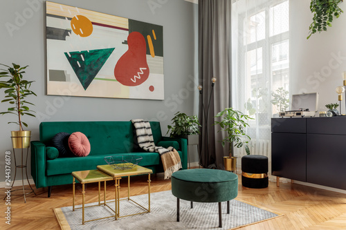 Luxury and modern home interior with design green sofa, navy commode, tables, pouf and accessroies. A lot of plants in the room. Abstract painting.  Stylish decor of living room with brown parquet. 