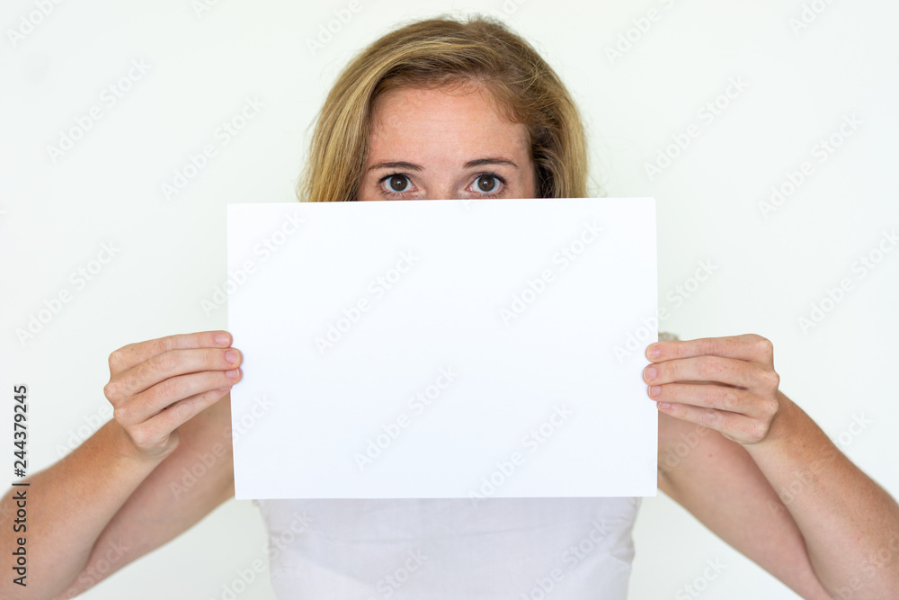 Young woman hiding behind blank sheet of paper. Lady looking at camera and advertising product. Promotion concept. Isolated front view on white background. Young woman hiding behind blank sheet of paper. Lady looking at camera and advertising product. Promotion concept. Isolated front view on white background.