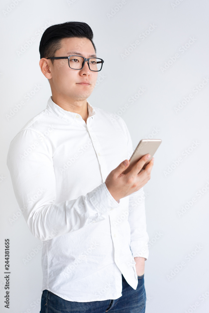 Pensive Asian man holding and using smartphone. Young guy standing. Technology concept. Isolated front view on white background. Pensive Asian man holding and using smartphone. Young guy standing. Technology concept. Isolated front view on white background.