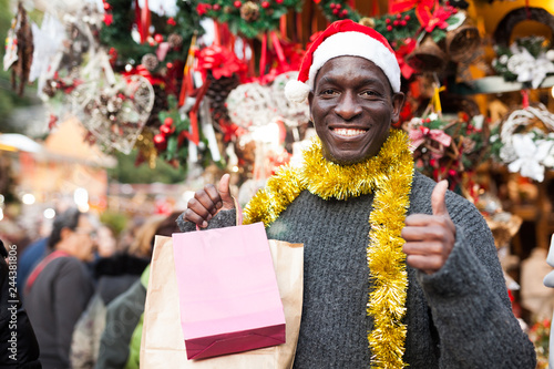 Smiling man in Santa hat with shopping bags on Christmas fair