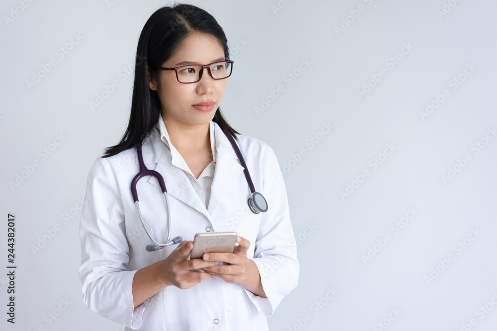 Serious young female doctor using smartphone. Pretty Asian woman wearing white coat and standing. Medicine and communication concept. Isolated front view on white background. Serious young female doctor using smartphone. Pretty Asian woman wearing white coat and standing. Medicine and communication concept. Isolated front view on white background.