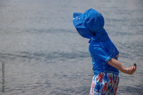 toddler boy wearing sun protective clothing and hat throws rock in ocean