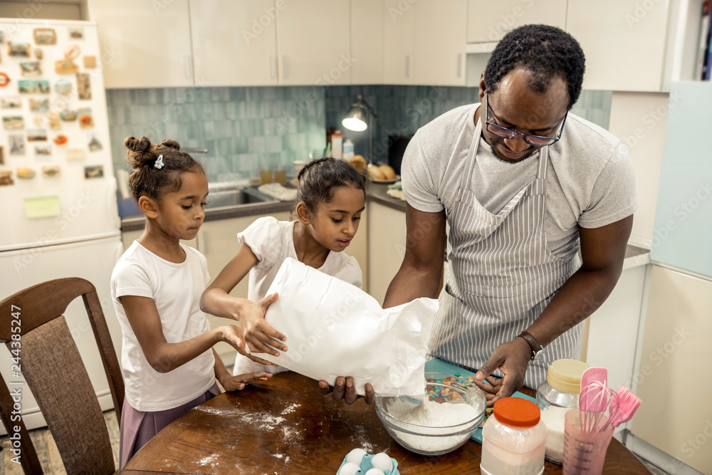 Father wearing glasses putting flour in bowl with daughters Father wearing glasses putting flour in bowl with daughters