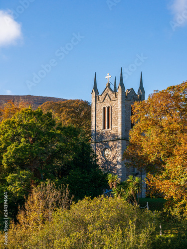 Irish church tower between autumn colored trees on a sunny day under a blue clear sky. St. Mochonog   s Church in Kilmacanogue Village  County Wicklow  Ireland.