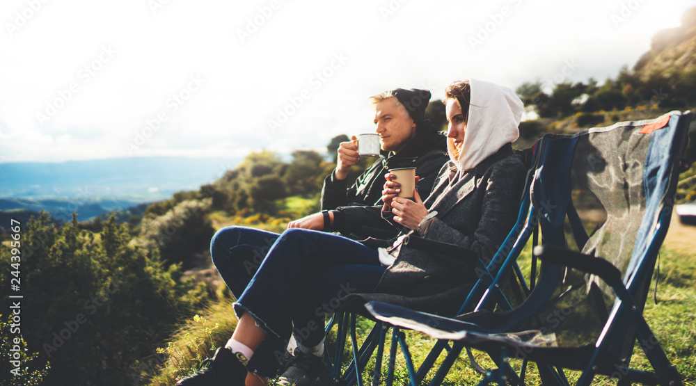 love couple sit in camping chairs on top of sun flare mountain, travelers drink tea on cup enjoy nature, romantic look into distance on background of panoramic landscape, weekend concept