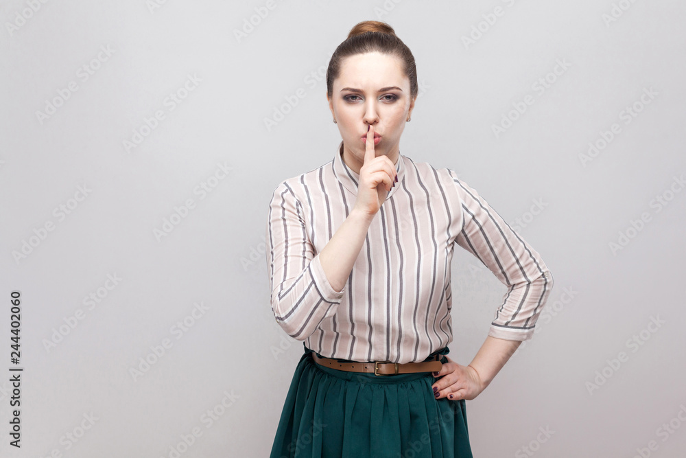 Fototapeta premium Be quiet! Serious beautiful young woman in striped shirt and green skirt with makeup and collected ban hairstyle, standing with finger on nose and silent sign. studio shot, isolated on grey background