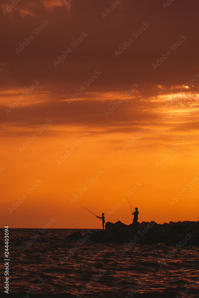 Two persons fishing with rods at seaside during sunset time. Vertical color photography.
