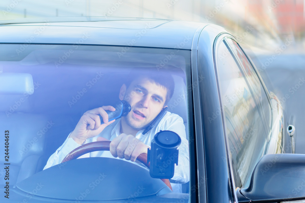 Naklejka premium A young guy is a black car driver talking on a mobile phone / smartphone, drinking tea, coffee and shaving with an electric razor. The concept of not safe driving