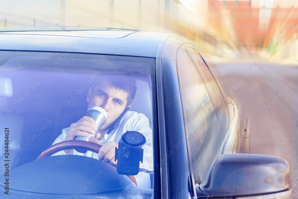 A young guy is a black car driver talking on a mobile phone / smartphone, drinking tea, coffee. The concept of not safe driving