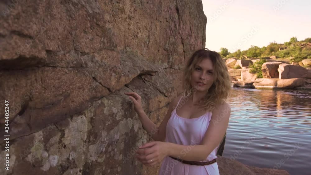 passionate and ambitious girl with blond blond curly hair plays on camera, spins smoothly and gracefully near high stone rock above the river, dances in long pink dress, laughs merrily and defiantly passionate and ambitious girl with blond blond curly hair plays on camera, spins smoothly and gracefully near high stone rock above the river, dances in long pink dress, laughs merrily and defiantly