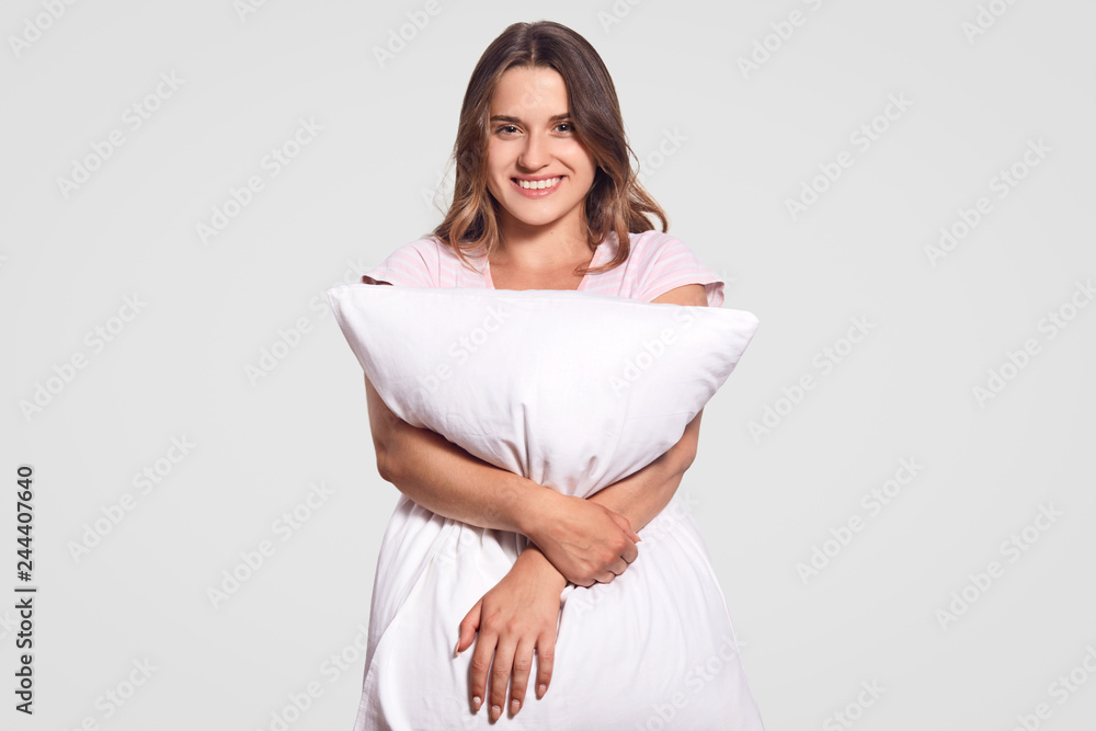 Image of positive Caucasian lady with toothy smile hugs pillow, wears pajamas, poses in studio against white background. Happy young woman in good mood after having pleasant dreams at night. Image of positive Caucasian lady with toothy smile hugs pillow, wears pajamas, poses in studio against white background. Happy young woman in good mood after having pleasant dreams at night.