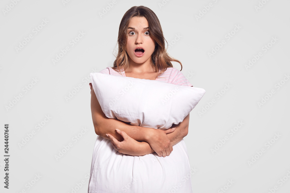 Astonished dark haired woman carries white soft pillow, horrified by nightmare, surprised by something terrible, keeps jaw dropped, looks with fear, isolated over white background. Sleeping concept Astonished dark haired woman carries white soft pillow, horrified by nightmare, surprised by something terrible, keeps jaw dropped, looks with fear, isolated over white background. Sleeping concept