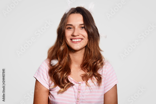 Bed time concept. Cheerful young woman with toothy smile, has feathers on head, wears pyjamas, ready for good rest, isolated over white background, expresses positive feelings, models indoor