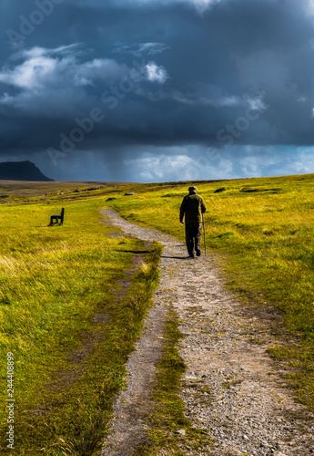Old Man With Walking Stick Strolls On Narrow Gravel Path Through Scenic Landscape With Spectacular Clouds