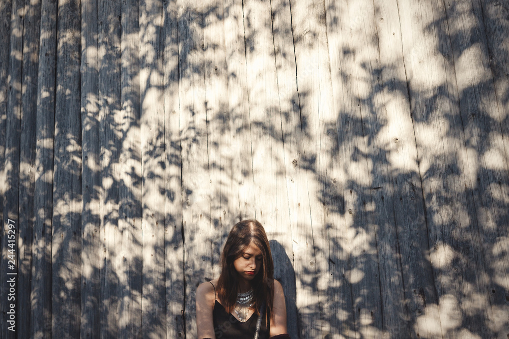 Stylish hipster girl posing in sunny street on background of wooden wall. Boho girl in cool outfit standing in sunlight and shadow. Space for text. Summer vacation and travel. Creative photo