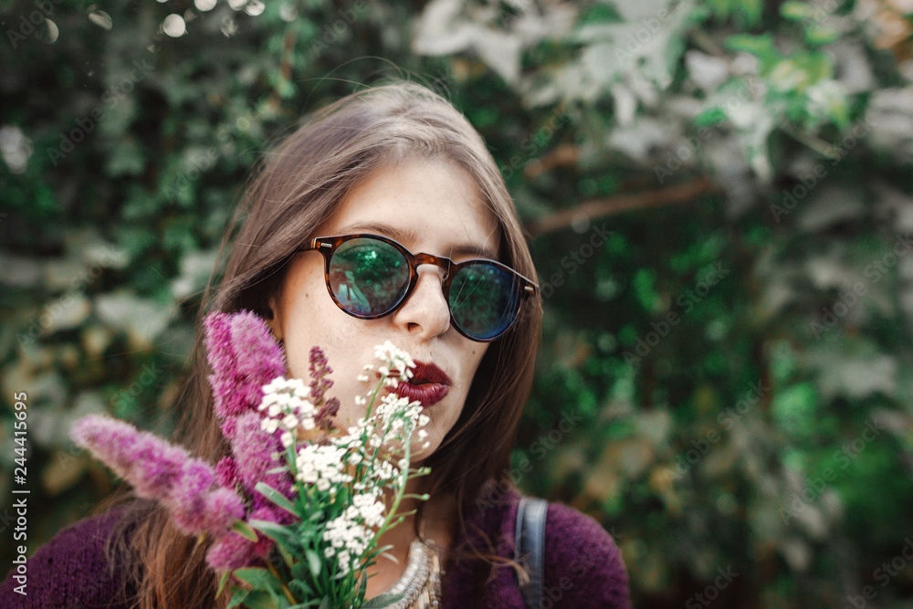 Portrait of happy boho girl in sunglasses smiling with bouquet of wildflowers in sunny garden. Stylish hipster carefree girl having fun at green bush in street. Summer vacation and travel Portrait of happy boho girl in sunglasses smiling with bouquet of wildflowers in sunny garden. Stylish hipster carefree girl having fun at green bush in street. Summer vacation and travel