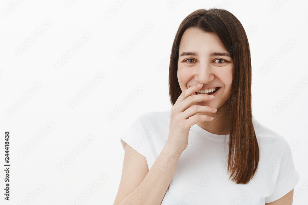Girl laughing over you as having fun and fooling around being in playful upbeat mood giggling, covering perfect smile with hand and gazing amused at camera posing in white t-shirt against gray wall Girl laughing over you as having fun and fooling around being in playful upbeat mood giggling, covering perfect smile with hand and gazing amused at camera posing in white t-shirt against gray wall