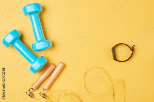 Flat lay composition of blue dumbbells, jump rope and fitness tracker on a yellow background with copy space © Lazy_Bear