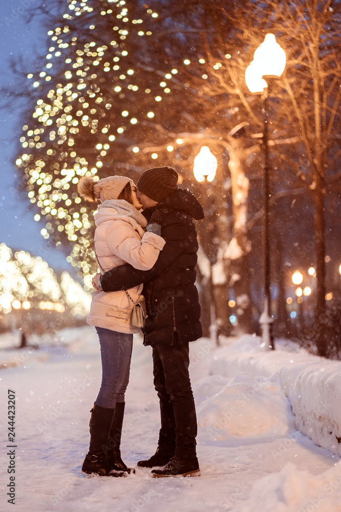 Young cheerful couple kiss, enjoying, dressed warm. Winter holidays, people concept - young couple in the evening walking under falling snow in park.